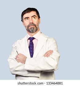Portrait of serious middle-aged dentist, looking at copyspace and thinking, wearing lab coat, dentist mirror in his hand on a pale background.