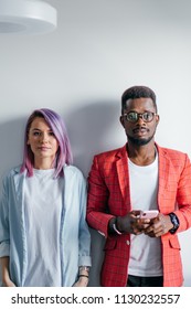 Portrait Of Serious No Emotion Face Multiracial Couple Isolated Over Gray Wall Background, Copy Space. African Man Dressed In Red Jacket And Caucasian Woman With Dyed Violet Hair Looking At Camera