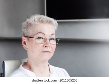 Portrait Of A Serious Older Woman In Glasses, Blonde With A Short Hairstyle, Aged, Sitting On A Chair In A Room And Looking At The Camera