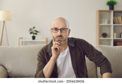 Portrait Of Serious Pensive Intelligent Man On Sofa At Home. Bald Adult Man In Glasses Sitting On Couch, Looking At Camera, Listening To Someone And Thinking. Video Call Laptop Computer Screen View