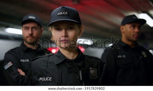 Portrait of serious\
policewoman in cap posing at camera with colleagues on background.\
Female cop standing at car with flashing lights. Police officer\
looking at camera 