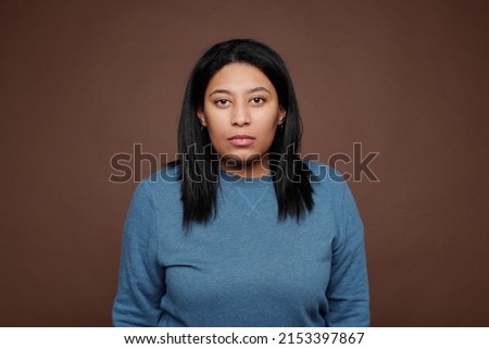 Portrait of serious portly Black woman in blue sweater standing against isolated brown background