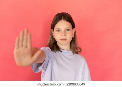 Portrait Of Serious Preteen Girl Making Stop Gesture. Caucasian Child Wearing Blue T-shirt Showing Palm Against Red Background. Home Violence And Childrens Rights Concept