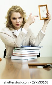 Portrait Of Serious Pretty Business Woman With Blonde Curly Hair In Jacket Showing On Sand Hour Glass Clock Sitting At Table With Heap Of Books On White Wall Background, Vertical Picture