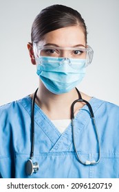 Portrait Of Serious Pretty Caucasian Young Female Doctor,wearing Blue Scrubs Uniform,protective Goggles,surgical Face Mask,stethoscope,isolated On White Background,UK NHS Hospital Or Clinic Staff