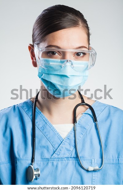 Portrait of serious pretty caucasian young female\
doctor,wearing blue scrubs uniform,protective goggles,surgical face\
mask,stethoscope,isolated on white background,UK NHS hospital or\
clinic staff