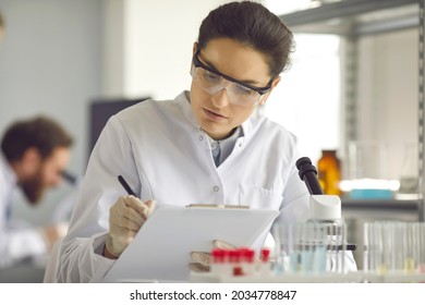 Portrait Of Serious Scientist In White Lab Coat Collecting Data In Medical Research Laboratory. Focused Med Student Holding Clipboard And Taking Notes Summarising Information For Scientific Report