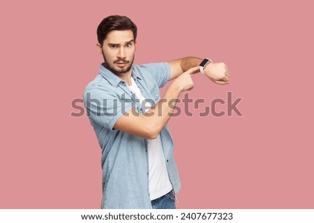 Portrait of serious strict bearded man in blue casual style shirt standing looking at camera, pointing at his smart watch, reminds about deadline. Indoor studio shot isolated on pink background.