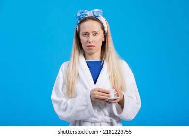 Portrait Of Serious Woman Holding Cup Of Coffee Wearing Bathrobe. The Housewife Mother Of Children Is Getting Ready For Work A Moment For Before Doing Chores. Blue Studio Background.