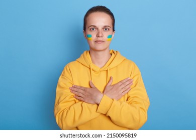 Portrait Of Serious Woman Wearing Hoodie With Blue And Yellow Flag On Cheeks, Isolated Over Blue Background, Keeps Hands Crossed On Her Chest, Looking At Camera. Glory To Ukraine.