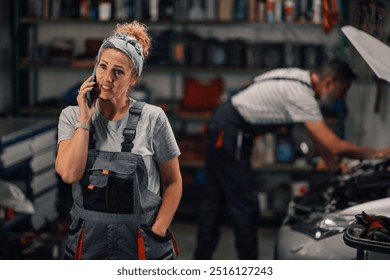 Portrait of serious worried female auto mechanic worker standing at mechanic workshop with cellphone in hand and talking on a phone with client. In a blurry background is male worker fixing automobile - Powered by Shutterstock