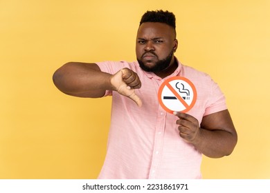 Portrait of serious young adult bearded man wearing pink shirt holding no smoking sign, showing thumb down gesture, looking at camera. Indoor studio shot isolated on yellow background. - Powered by Shutterstock