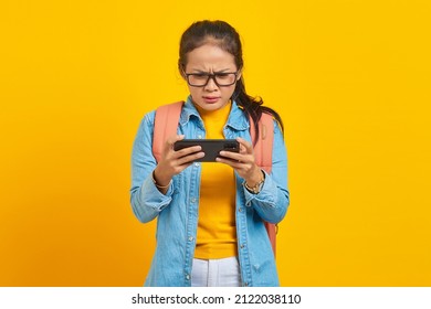 Portrait Of Serious Young Asian Woman Student In Casual Clothes With Backpack Playing Video Game On Mobile Phone Isolated On Yellow Background.  Education In College University Concept