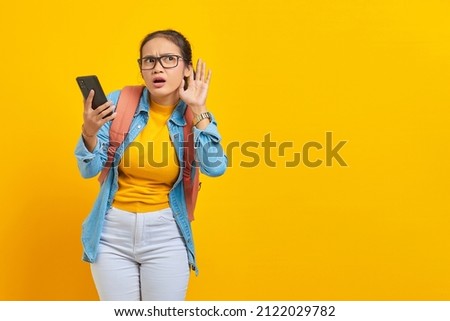 Portrait of serious young Asian woman student in casual clothes with backpack holding smartphone and trying to overhear secret conversation isolated on yellow background