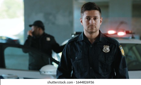 Portrait serious young man cops stand near patrol car look at camera enforcement background his colleague happy officer police uniform auto safety control policeman close up slow motion