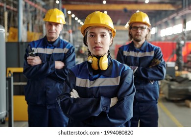 Portrait of serious young woman in hardhat and noise-cancelling headphones working with male colleagues at factory