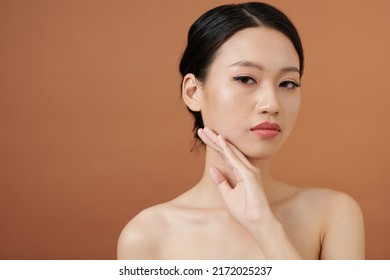 Portrait Of Serious Young Woman Touching Face And Looking At Camera, Isolated On Brown Background