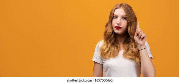 Portrait Of Serious Young Woman With Wavy Redhead Showing Index Finger, Scolding Or Telling Off Someone. Girl Raises Finger Up, Says: Attention Please. Copy Space For Your Text