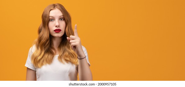 Portrait Of Serious Young Woman With Wavy Redhead Showing Index Finger, Scolding Or Telling Off Someone. Girl Raises Finger Up, Says: Attention Please. Copy Space For Your Text