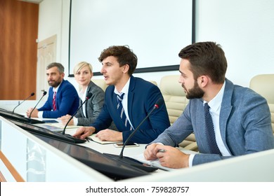 Portrait Of Several Business People Sitting In Row Participating In Political Debate During Press Conference Answering Media Questions Speaking To Microphone