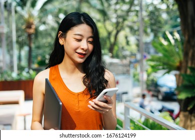 Portrait Of Sexy Asian Beautiful Woman Wearing Orange Shirt Holding Laptop, Talking On The Phone At Cafe Garden In The City.