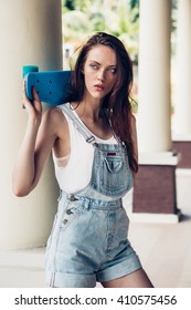 Portrait Of Sexy Beautiful Woman In White T-shirt And Short Jeans Overall Holding Blue Short Skate Board. Urban Scene, City Life. Cute Attractive Sexy Hipster Lady Standing In Front Of A Column.