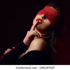 Portrait of Sexy brunette pretty woman model in black clothes and red transparent mask on eyes touching lips over dark background. Stylish look and sexual games concept
