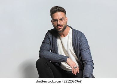 Portrait Of Sexy Casual Man In Hoodie With Glasses Holding Elbow On Knee And Crouching In Front Of Grey Background In Studio