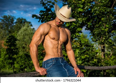 Portrait of sexy farmer or cowboy in hat looking to a side, while standing next to hay field in countryside