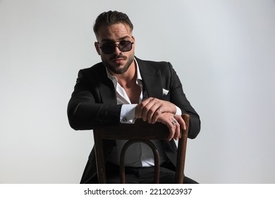 Portrait Of Sexy Man With Open Collar Shirt And Sunglasses Holding Arms On Chair And Posing On Grey Background In Studio