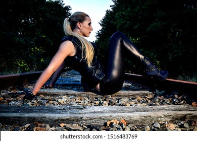 Portrait Of A Sexy Woman In Black Leather Clothes And Gloves. Gangsta Girl With Red Lips And Blonde Hair Sitting On The Abandoned Railway. 