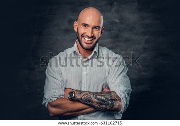 Portrait of shaved head male in a white shirt with\
tattooed crossed arms.