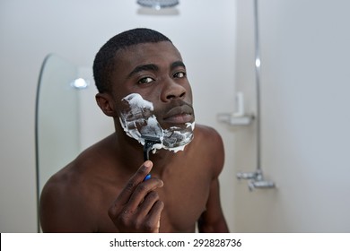 Portrait Of Shirtless African Black Man Shaving Beard Stubble Face With Razor In Mirror Reflection For Morning Clean Shaven Look In Home Bathroom