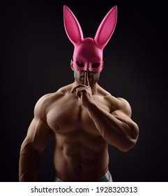 Portrait of shirtless handsome muscular man, athlete with perfect built body wearing pink rabbit, bunny mask stands with naked chest and gesturing silence, secret sign with finger on dark background