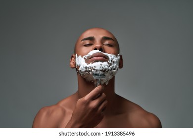 Portrait of shirtless young african american man using steel razor for removing stubble from neck, posing with eyes closed isolated over gray background. Horizontal shot - Powered by Shutterstock