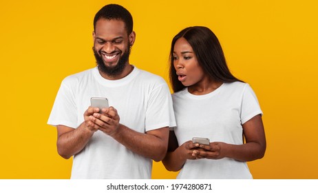 Portrait of shocked African American woman spying on her smiling boyfriend who using mobile phone, texting sms or scrolling social media news feed. Black couple standing over yellow studio background