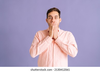 Portrait of shocked amazed excited young man standing covering mouth with hands looking camera on isolated purple background in studio. Scared guy hocked covering mouth with hands for mistake.