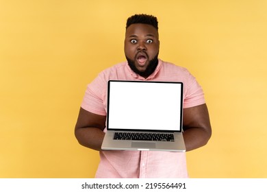 Portrait of shocked amazed man wearing pink shirt holding notebook, showing white blank screen on his laptop, looking at camera with big eyes. Indoor studio shot isolated on yellow background.