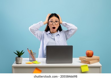 Portrait Of Shocked Asian Female Student Sitting At Desk With Laptop Computer And Grabbing Her Head Isolated Over Blue Studio Background. Stressed Lady Studying And Reading News