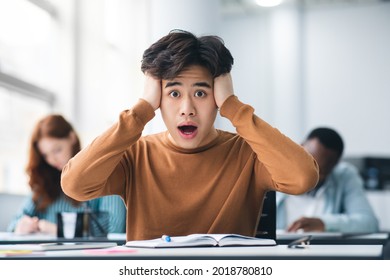 Portrait of shocked asian male student sitting at desk in classroom, grabbing his head looking at camera. Worried youth unprepared for test or exam, thinking about deadline or hard new theme