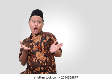 Portrait of shocked Asian man wearing batik shirt and songkok shouting wow with pointing hands with open palms gesture. Isolated image on gray background