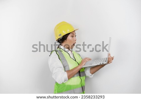 A portrait of a shocked Asian woman labor wearing safety helmet and vest while holding her laptop with her mouth wide open, isolated by white background. Labor's day concept