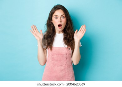 Portrait of shocked attractive woman gasping, spread hands sideways and stare startled at camera, hear worrying terrible news, looking with disbelief, standing against bue background