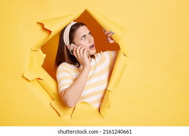 Portrait Of Shocked Attractive Woman Stands In Torn Paper Hole And Looking Away With Open Mouth While Talking Phone, Looking Through Breakthrough Of Yellow Background.