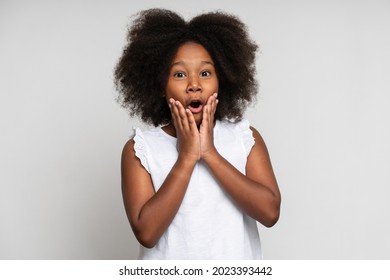 Portrait of shocked beautiful brunette young girl with black curly hair in white dress standing and looking at camera with surprised face. Indoor studio shot isolated on white background 