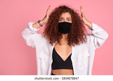 Portrait Of Shocked Ginger Girl With Curly Hair, Wearing A Medical Mask To Protect Against Coronavirus. Coronavirus Causes Covid-19 Disease Isolated Over Pink Background