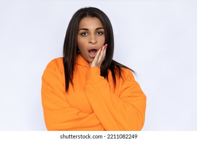 Portrait Of Shocked Hispanic Woman With Open Mouth. Surprised Young Model With Long Hair In Orange Hoodie Looking At Camera, Touching Face. Rumor, Secret Concept.