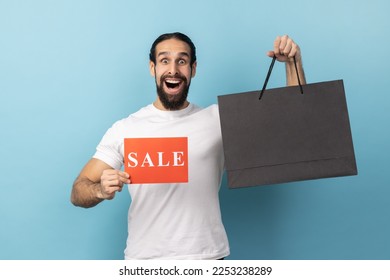 Portrait of shocked man with beard wearing white T-shirt holding shopping bags and showing sale card, advertising low prices in the mall. Indoor studio shot isolated on blue background. - Shutterstock ID 2253238289