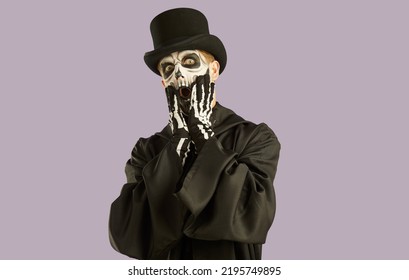 Portrait of shocked man in Halloween make-up and costume grabbing his face in fright. Man in black hat, suit and skull make-up opens his eyes and mouth wide in fear on light lilac background.