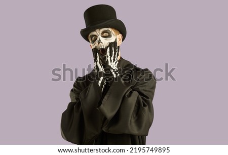 Portrait of shocked man in Halloween make-up and costume grabbing his face in fright. Man in black hat, suit and skull make-up opens his eyes and mouth wide in fear on light lilac background.